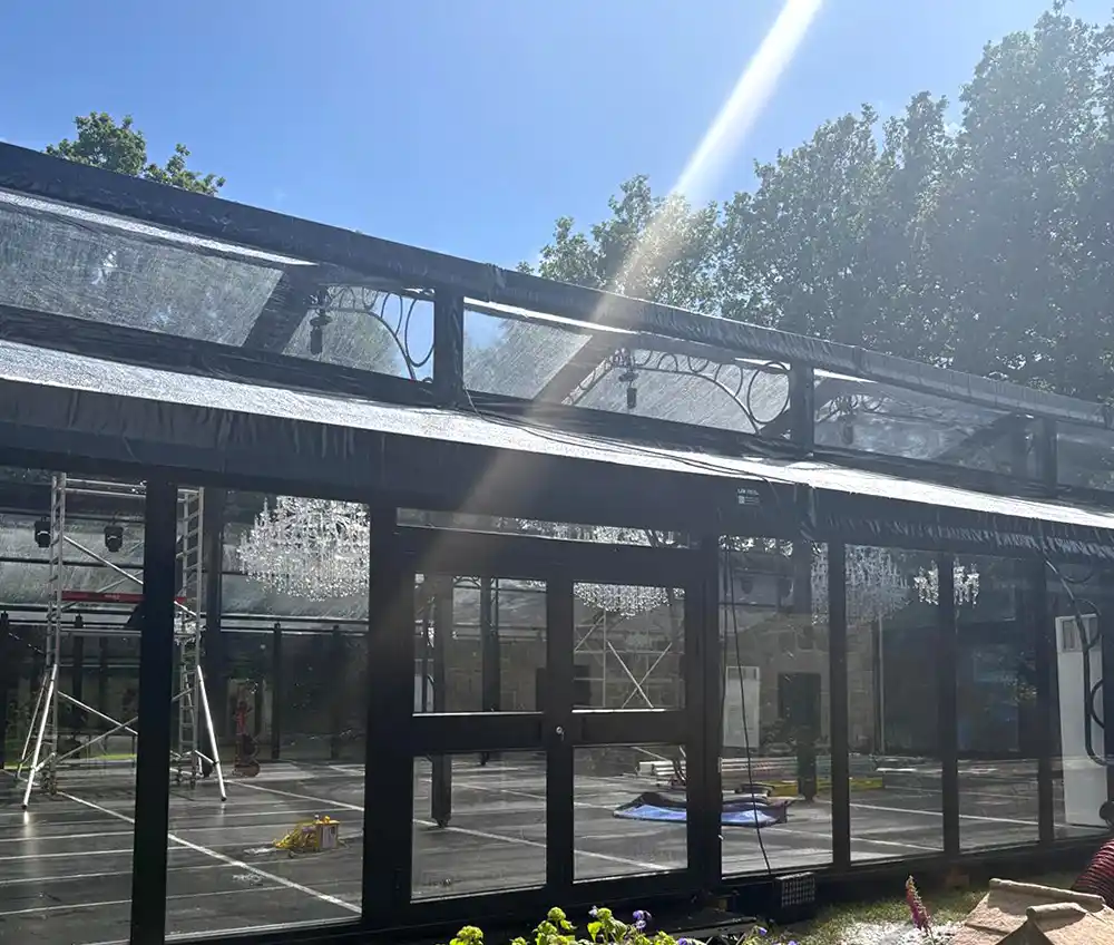 Glass Wedding Marquee Structure Ahead of Event Setup Glass-fronted wedding marquee with floor-to-ceiling glazing and hanging chandeliers, photographed during setup ahead of a summer wedding